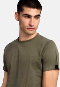 Tricou din bumbac de culoare verde olive cu guler rotund, mâneci scurte și un eticheta neagră cuvântul "REPLAY" pe mâneca stângă.