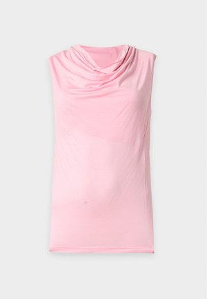 Vrouw staand, gekleed in een mouwloze roze top, losse witte broek en zwarte schoenen tegen een effen lichte achtergrond.
