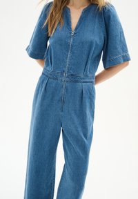 Denim-Overall mit kurzen Ärmeln und V-Ausschnitt, mit einem Frontreißverschluss, tailliertem Schnitt und weiten Beinen in einem mittleren Blauton.