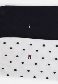 Due paia di calzini: uno nero uniforme e uno bianco con pois neri. Entrambi presentano un piccolo logo ricamato con accenti rossi, bianchi e blu.
