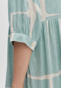 Hellblau gemustertes Kleid mit schichtigen Design, das über Dreiviertelärmel, einen geknöpften Bund und einen strukturierten Stoff verfügt.