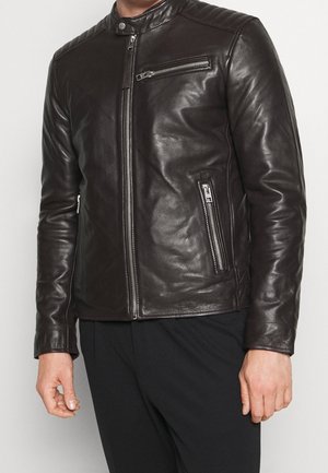 Mężczyzna noszący dopasowaną ciemnobrązową skórzaną kurtkę z zamkami błyskawicznymi na kieszeniach na piersi i po bokach, stojący na jednolitym tle.