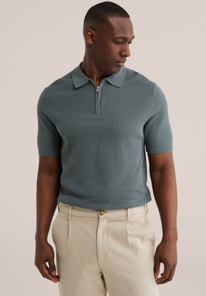 Muž v krátkym rukávom, textúrovanom sivozelenom polokošeli na zips a béžových plisovaných nohaviciach, pozerajúci sa dole na ľavú stranu.