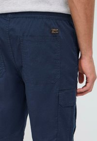 Mørkeblå cargo shorts med flere lommer, et teksturert stoff og et logo-merke nær midjen. Har en avslappet passform.