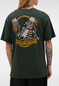 Mężczyzna z tatuażami, ubrany w ciemnozieloną koszulę z grafiką czaszki w futrzanym kapeluszu trzymającą mrożony kufel piwa, z napisem "Vans Off The Wall Freezing Cold Since 1966".