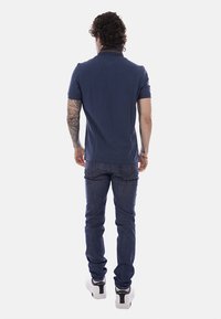 Homme aux cheveux bouclés et tatouages, portant un polo marine, un jean bleu foncé et des baskets blanches, debout de dos sur fond blanc.