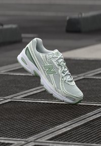 Hele rohelised ja valged New Balance jooksujalatsid, mis hõljuvad linna keskkonnas metallist tänavakanalite kohal.