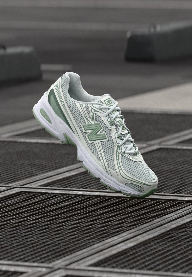 Hele rohelised ja valged New Balance jooksujalatsid, mis hõljuvad linna keskkonnas metallist tänavakanalite kohal.