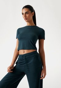 Donker teal sportieve cropped top met korte mouwen en geborduurd logo; gecombineerd met bijpassende losse broek met elastische tailleband.