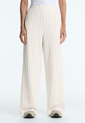 Kobieta ubrana w kremowe spodnie z wysokim stanem i szerokimi nogawkami z cienkimi pionowymi czarnymi paskami oraz białe trampki, stojąca na tle jednolitego tła.