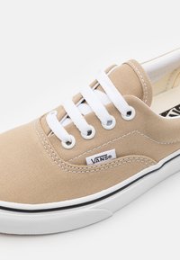 Béžová teniska Vans s bílými tkaničkami, bílou stélkou, černým pruhem a štítkem s logem Vans na boku.