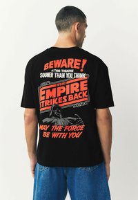 Tricou din bumbac negru cu un text grafic îndrăzneț în roșu și alb și imagini din "Star Wars: The Empire Strikes Back" pe spate.