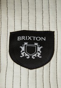 Patch logo Brixton sur un tissu crème avec des rayures noires. La forme du bouclier présente un fond noir texturé avec des lettres et des accents en argent.