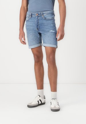 Helle blaue Denim-Shorts mit fransigem Saum, umgeschlagenen Bündchen und subtilen Gebrauchsspuren. Kombiniert mit weißen und grauen Turnschuhen sowie gerippten Socken.