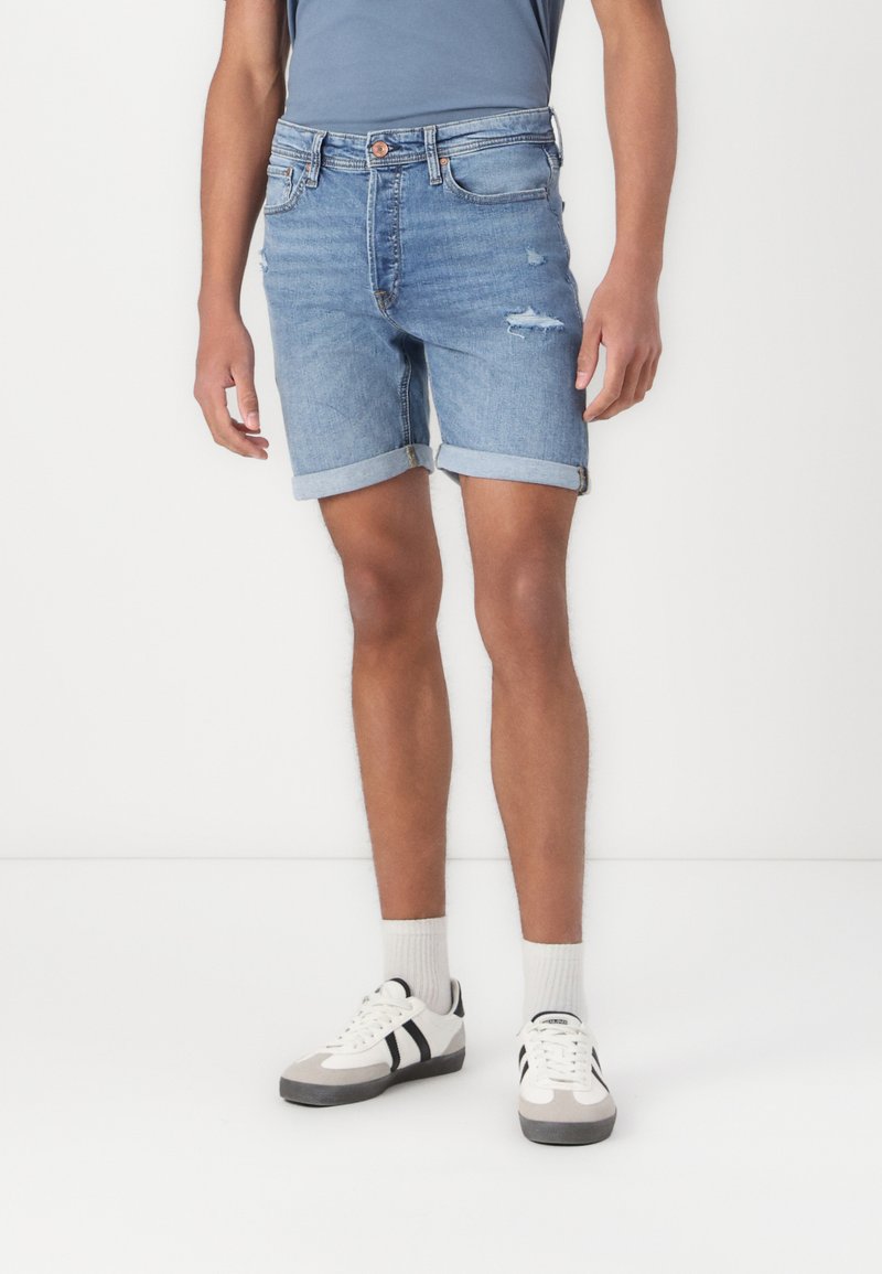Shorts en denim bleu clair avec un ourlet effiloché, des poignets roulés et une légère décoloration. Associés à des baskets blanches et grises et des chaussettes côtelées.