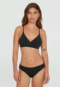 Sieviete ar gariem brūniem matiem un tetovējumiem, tērpta melnā bikini komplektā, stāv uz vienkrāsainas gaišas fona, vērsta uz priekšu.