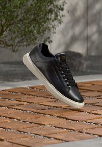 Schwarzer Ledersneaker mit weißer Gummisohle, ausgestattet mit Schnürverschluss und goldfarbenen Logoakzenten an der Seite und auf der Zunge. Glatte Textur.