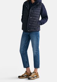 Svart puffig väst över en marinblå hoodie, blå kortbyxor och bruna sneakers med nätdetaljer. Tryckknappar och sidofickor på västen.