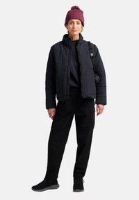 Giacca nera trapuntata con colletto alto, camicia scura, pantaloni cargo neri e scarpe nere. Indossata con un berretto lavorato in marezzato, con pompon.