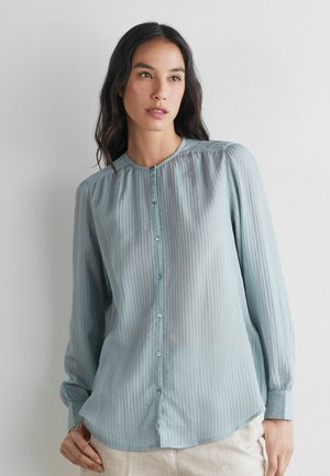 Femme portant un chemisier bleu clair à manches longues, boutonné, avec des rayures verticales, debout devant un mur blanc uni.