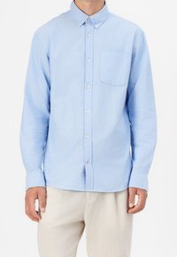 Camisa de botones azul claro con mangas largas, un bolsillo en el pecho y dobladillo redondeado, hecha de una tela suave y texturizada. Presenta botones blancos.