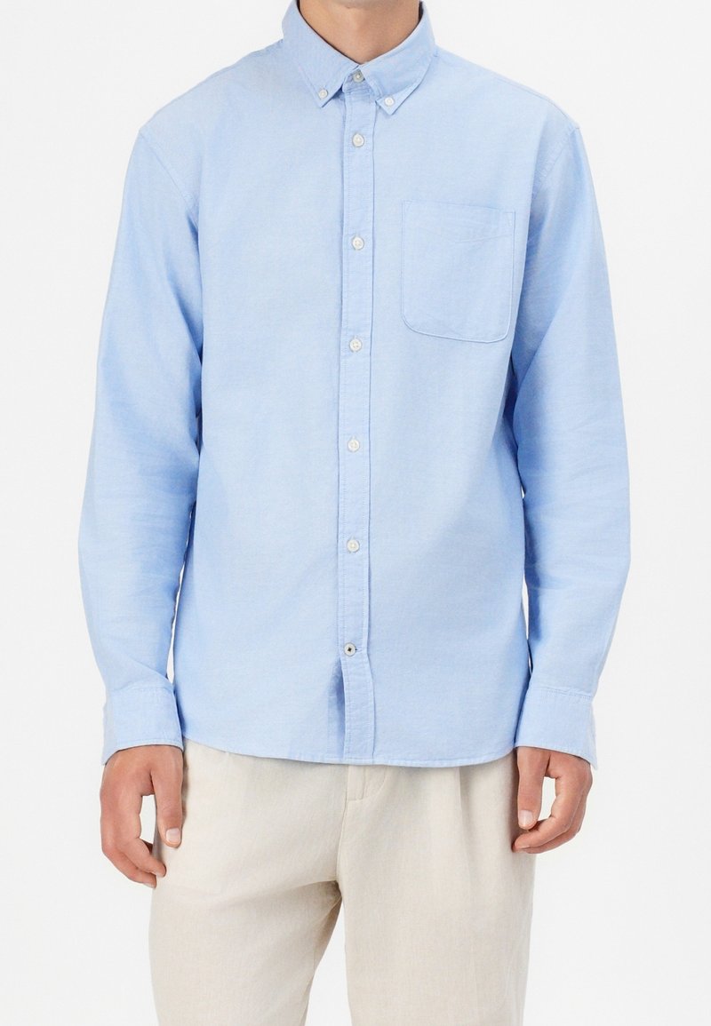 Camisa de botones azul claro con mangas largas, un bolsillo en el pecho y dobladillo redondeado, hecha de una tela suave y texturizada. Presenta botones blancos.