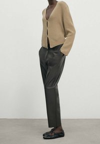 Beiger Strickcardigan mit tiefem V-Ausschnitt und vorderen Knöpfen, kombiniert mit eng anliegenden dunklen Lederhosen und schwarzen Schnürschuhen.