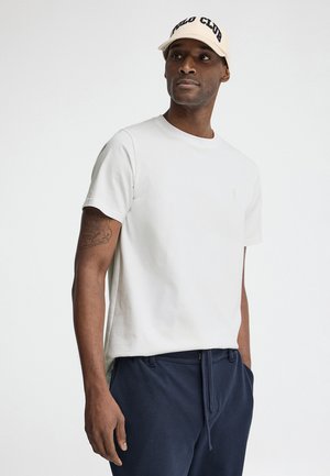 Mężczyzna ubrany w kremowy T-shirt, granatowe spodnie i beżową czapkę z napisem „POLO CLUB”, stoi na tle jednolitego jasnego tła.