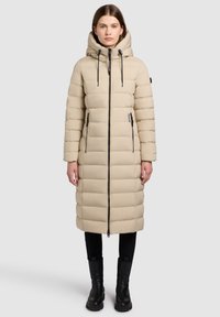 Manteau matelassé beige avec capuche, doté d'une fermeture éclair noire et de cordons de serrage, poches latérales et design long matelassé. Leggings noirs et bottes.