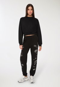 Młoda kobieta ubrana w czarną, krótką bluzę z długim rękawem, czarne dresy z białym graffiti oraz srebrne sneakersy, stojąca na białym tle.