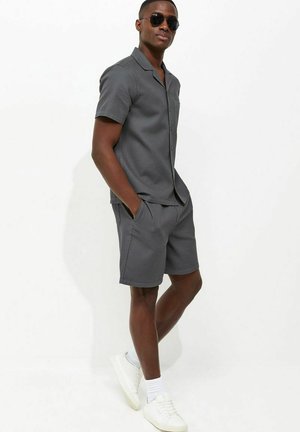 Mężczyzna w ciemnoszarej koszulce z krótkim rękawem i pasujących szortach, białe trampki, okulary przeciwsłoneczne, stoi z rękami w kieszeniach na białym tle.