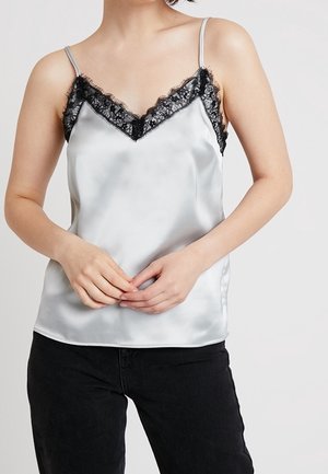 Mujer con camiseta de satén plateado con bordes de encaje negro y tirantes finos, combinada con pantalones negros, de pie frente a un fondo claro.