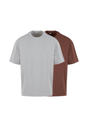 Jeune homme aux cheveux courts portant un t-shirt large gris clair uni et un pantalon noir, debout les mains dans les poches, regardant sur le côté.