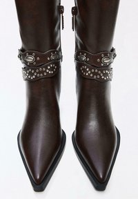 Ciemnobrązowe skórzane kozaki z ostro zakończonym noskiem, ozdobione srebrnymi ćwiekami i owalnymi metalowymi elementami na paskach wokół kostek.