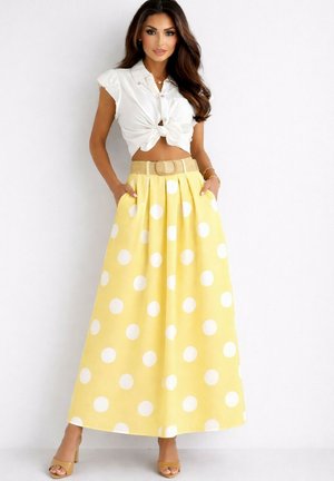 Femme portant un crop top blanc noué, une jupe longue jaune taille haute à pois blancs, une large ceinture beige et des talons beiges, debout contre un mur blanc.