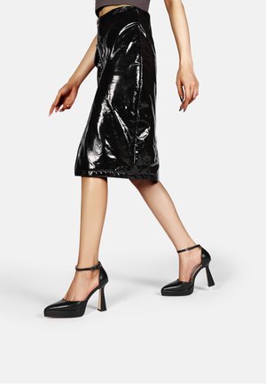 Tacchi a piattaforma neri con una finitura lucida e elegante, cinturino alla caviglia e un alto tacco a blocco, abbinati a una gonna a tubo nera lucida.