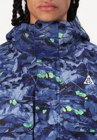 Blue camouflage jacket with a glossy finish, featuring green leaf patterns, a hood, and a small triangular logo on the chest.