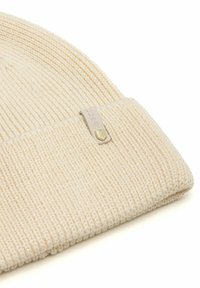 Beżowy, prążkowany czapka typu beanie w kolorze kremowym z podwinięciem i małą prostokątną beżową metką z złotym heksagonalnym emblematem.