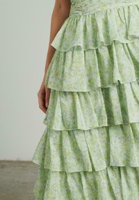 Jasnozielona sukienka warstwowa z falbanami, ozdobiona kwiatowym wzorem. Tkanina jest miękka i lekko przezroczysta, odsłaniając subtelne tekstury.