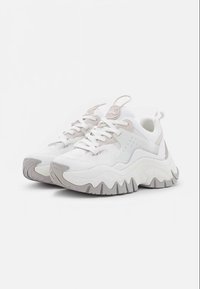 Zapatillas deportivas gruesas en blanco y gris claro con suelas gruesas onduladas, detalles de malla y cuero, y cierre de cordones, colocadas en ángulo sobre un fondo blanco liso.