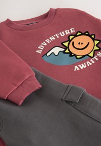 Bordo spalvos džemperis su saulės grafika ir užrašu „ADVENTURE AWAITS“, derinamas su pilkomis sportinėmis kelnėmis. Ribojuotos rankogaliai ir minkštas tekstūros audinys.