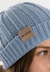 Jasnoniebieska dziana czapka z prążkowanym wzorem i beżową naszywką z napisem "FABRYKA DZIANIN" przyszytą do pasa. Miękka tekstura.