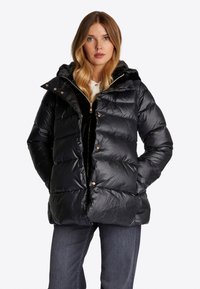 Schwarze Daunenjacke mit glänzendem Finish, horizontalem Steppmuster, Kapuze und Details aus goldfarbenem Hardware; über einem schwarzen Reißverschlussoberteil getragen.