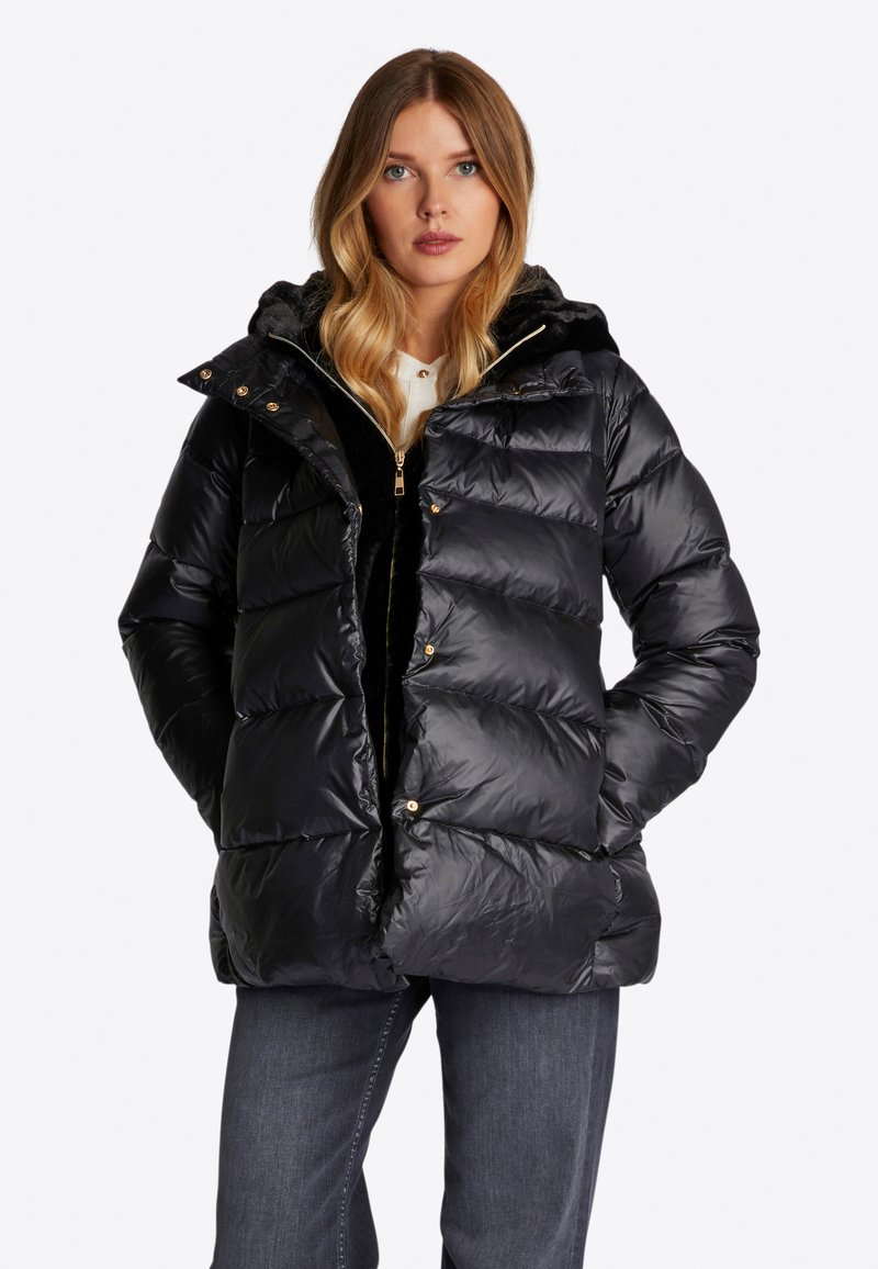 Schwarze Daunenjacke mit glänzendem Finish, horizontalem Steppmuster, Kapuze und Details aus goldfarbenem Hardware; über einem schwarzen Reißverschlussoberteil getragen.