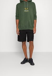 Zelená mikina s grafikou motýla a textem, spárovaná s černými šortkami. Outfit doplňují černé tenisky.