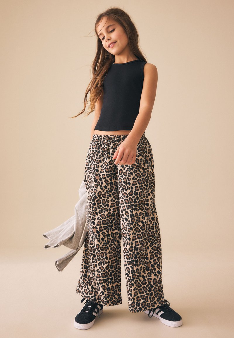 Fille portant un haut noir sans manches, un large pantalon imprimé léopard, des baskets noires et blanches, tenant une veste grise sur un fond beige.