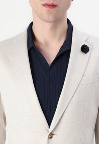 Lys beige blazer med teksturert stoff, enkel knappelukking, og en marineblå, ribbet V-hals skjorte under. Har en marineblå stoffblomst nål.