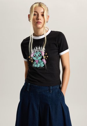 Jonge vrouw met lang blond haar draagt een zwart ringer T-shirt met een turquoise en roze cartoonhondenafbeelding, gecombineerd met een donkerblauwe plooirok.