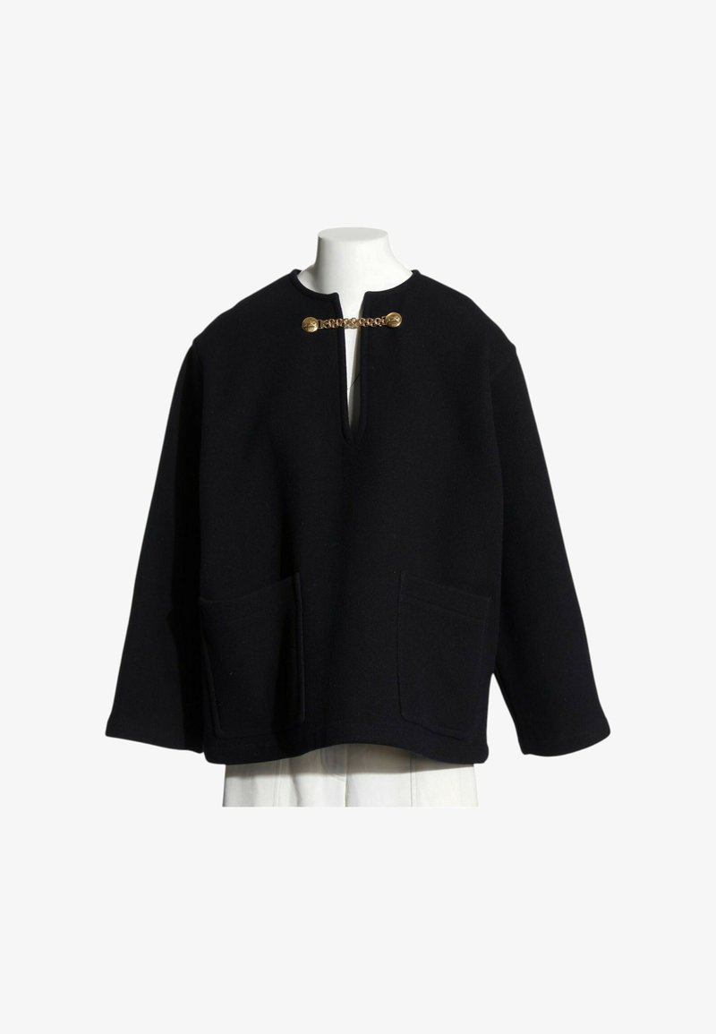 Veste noire à manches longues avec poches plaquées devant et fermoir en chaîne dorée au col ouvert, exposée sur un buste de mannequin blanc.