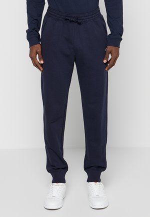 Uomo che indossa pantaloni della tuta blu navy con elastico in vita e coulisse, abbinati a sneaker bianche e una maglia a maniche lunghe blu navy.